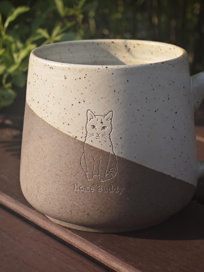 May include: A two-toned ceramic mug featuring a speckled cream upper section and a solid brown lower section. An embossed illustration of a seated cat is centred on the front, positioned above the engraved text Home Buddy in a simple font.
