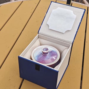 May include: A ceramic bowl with a lid, featuring a purple and blue glaze, is presented inside a navy blue box with a white interior. The box is open, revealing the bowl nestled within a soft, white cushion.
