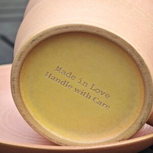 May include: Close-up of the bottom of a peach-colored ceramic mug with a yellow base. The text "Made in Love Handle with Care" is etched into the base. A pink plate is visible in the foreground.