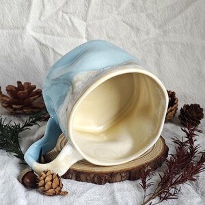 May include: A handmade ceramic mug with a light blue and white marbled exterior and a cream-colored interior. The mug has a curved handle and sits on a wooden slice, with pinecones and greenery around it.