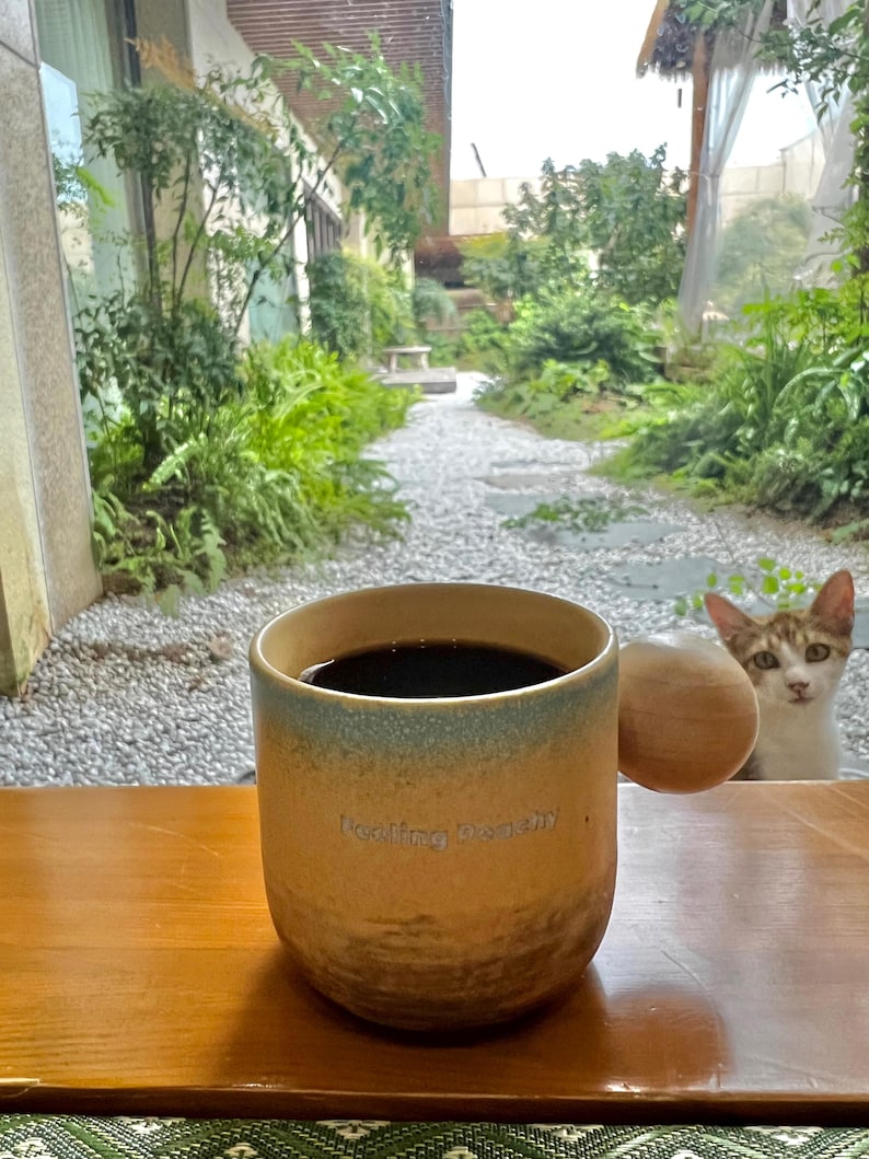 May include: A ceramic mug filled with dark liquid, possibly coffee, sits on a wooden tray. The mug has a tan base with a blue-green rim and a wooden handle. The text "Feeling Dandy" is printed on the mug. A cat is visible in the background.