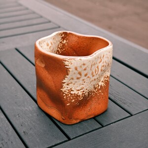 Peut inclure: Un vase en c&eacute;ramique fait &agrave; la main avec un design textur&eacute; unique. Le vase a une forme carr&eacute;e avec un bord sup&eacute;rieur ondul&eacute;. L'ext&eacute;rieur est brun terreux chaud, contrast&eacute; par un motif blanc tachet&eacute; sur la moiti&eacute; sup&eacute;rieure. Le vase est pos&eacute; sur une surface en bois gris fonc&eacute;.