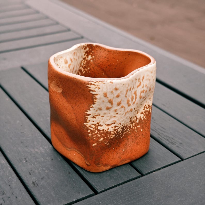 Peut inclure: Un vase en c&eacute;ramique fait &agrave; la main avec un design textur&eacute; unique. Le vase a une forme carr&eacute;e avec un bord sup&eacute;rieur ondul&eacute;. L'ext&eacute;rieur est brun terreux chaud, contrast&eacute; par un motif blanc tachet&eacute; sur la moiti&eacute; sup&eacute;rieure. Le vase est pos&eacute; sur une surface en bois gris fonc&eacute;.