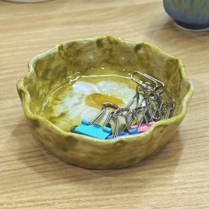 May include: A small, scalloped-edge ceramic bowl in shades of olive green and yellow, featuring a painted daisy design. The bowl contains several silver binder clips with blue and pink handles. The bowl sits on a light-colored wooden surface.