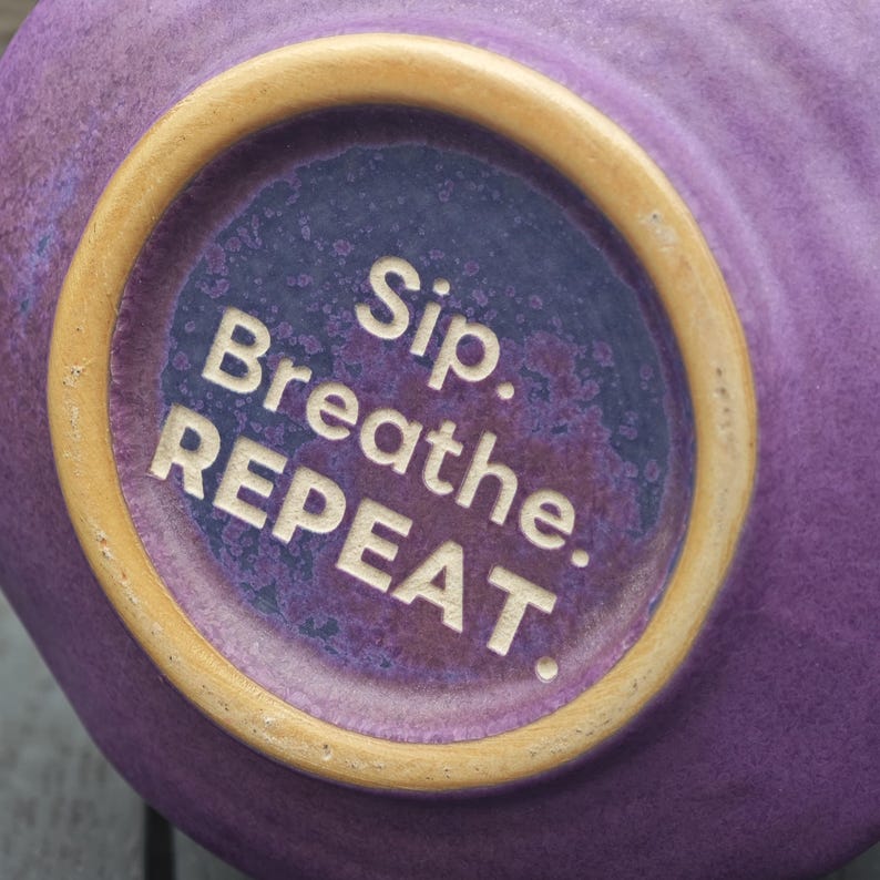 Op de afbeelding: Close-up van de bodem van een paarse keramische mok met een lichtbruine rand. Het midden van de bodem heeft de woorden "Sip. Breathe. REPEAT." in witte letters. De mok heeft een getextureerd oppervlak.