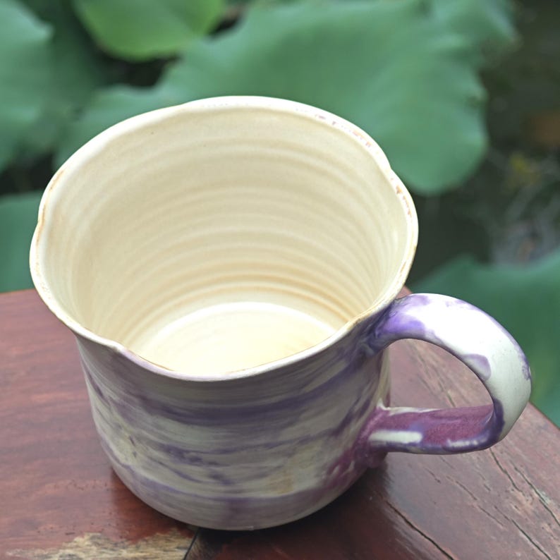 May include: Handmade ceramic mug with a unique, wavy rim. The mug features a cream-colored interior and a purple and white marbled exterior. The handle is also purple and white. A rustic, artistic piece for enjoying beverages.