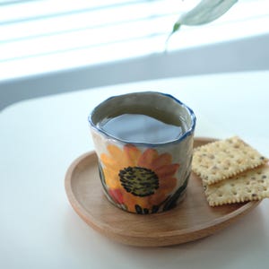 May include: A ceramic mug filled with tea, featuring a hand-painted sunflower design in orange, yellow, and red. The mug sits on a wooden tray with two crackers. The rim of the mug is blue.