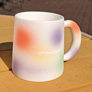 May include: White ceramic mug with a colorful gradient design featuring orange, purple, yellow, and green spots. The mug has a rounded handle and the text "off the hook" in white.