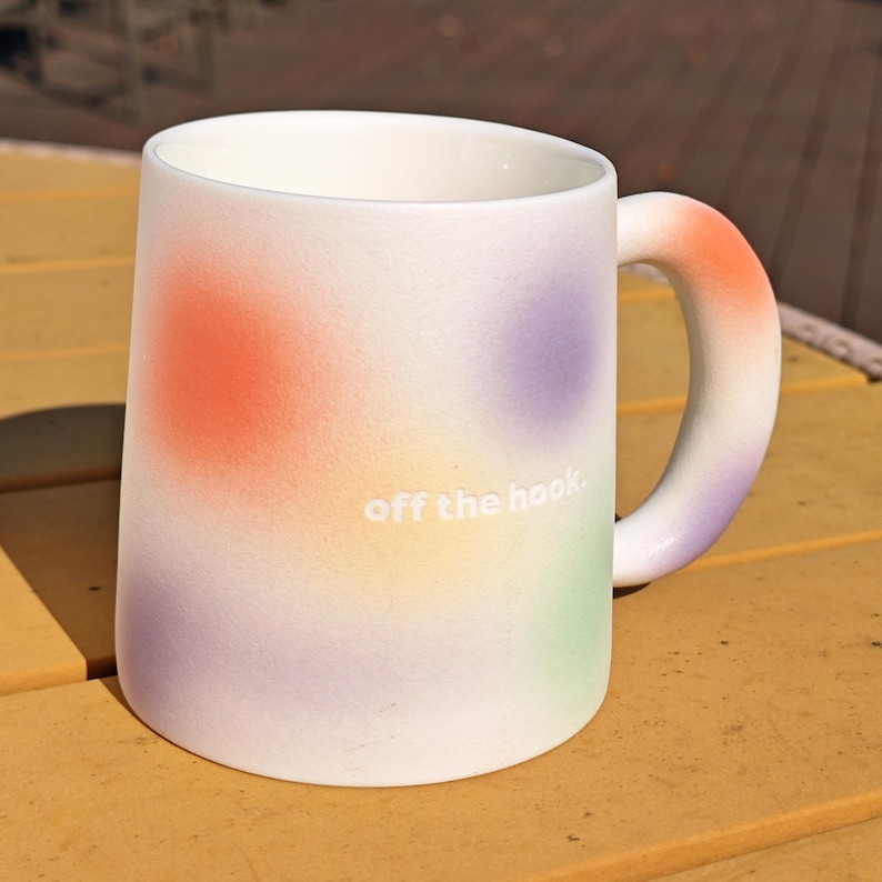 K&ouml;nnte beinhalten: Wei&szlig;er Keramikbecher mit einem farbenfrohen Farbverlauf-Design mit orangefarbenen, lila, gelben und gr&uuml;nen Flecken. Der Becher hat einen abgerundeten Griff und den Text "off the hook" in Wei&szlig;.