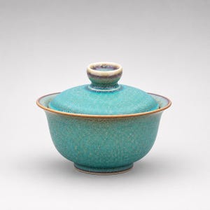 K&ouml;nnte beinhalten: Eine t&uuml;rkisfarbene Gaiwan-Teeschale aus Keramik mit passendem Deckel. Das Gef&auml;&szlig; verf&uuml;gt &uuml;ber eine gesprenkelte, texturierte Glasur mit subtilen violetten und braunen Verl&auml;ufen am Rand und am Knauf, auf einer abgerundeten Basis mit ausgestelltem Rand.