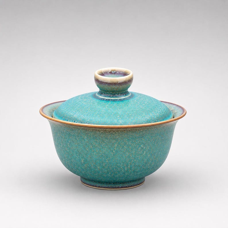 K&ouml;nnte beinhalten: Eine t&uuml;rkisfarbene Gaiwan-Teeschale aus Keramik mit passendem Deckel. Das Gef&auml;&szlig; verf&uuml;gt &uuml;ber eine gesprenkelte, texturierte Glasur mit subtilen violetten und braunen Verl&auml;ufen am Rand und am Knauf, auf einer abgerundeten Basis mit ausgestelltem Rand.