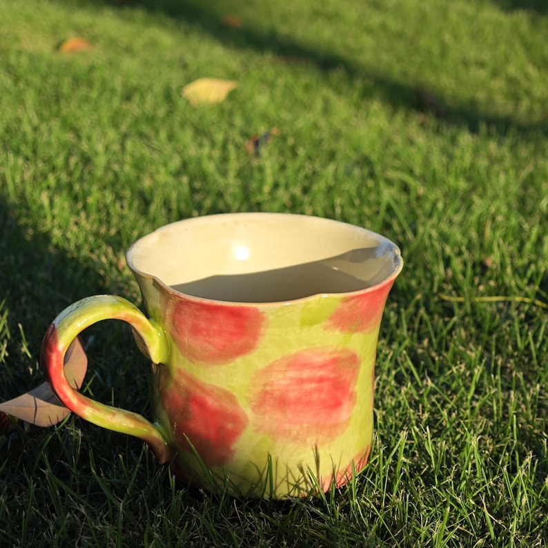 May include: A handmade ceramic mug with a unique design. The mug features a light green base adorned with large red polka dots. The handle and rim are painted in a complementary green. The interior is a creamy off-white color. The mug is sitting on a bed of green grass.