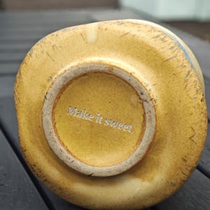 Peut inclure: Un bol en c&eacute;ramique jaune &agrave; la forme irr&eacute;guli&egrave;re. Le fond du bol porte l'inscription "Make it sweet" en lettres blanches. Le bol a un aspect rustique et fait main.