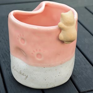 May include: A pink ceramic mug with a white speckled base and a small cat figurine attached. The mug features paw prints and the word "Lowkey" in cursive. The mug is designed with a unique curved top.