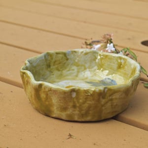 May include: A handmade ceramic bowl with a wavy rim. The bowl has a mottled, earthy color palette of olive green, tan, and hints of blue. The exterior has a textured appearance, suggesting a rustic, artisanal quality. A few small flowers are in the background.