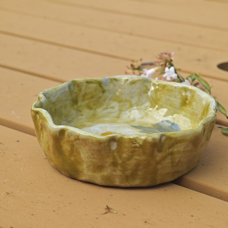 May include: A handmade ceramic bowl with a wavy rim. The bowl has a mottled, earthy color palette of olive green, tan, and hints of blue. The exterior has a textured appearance, suggesting a rustic, artisanal quality. A few small flowers are in the background.