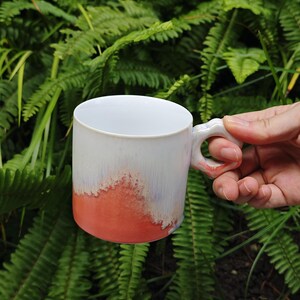 May include: White ceramic mug with a unique handle design. The mug features a gradient of colors, transitioning from white at the top to a vibrant coral at the base. The handle is a distinctive shape, adding to the mug's artistic appeal.