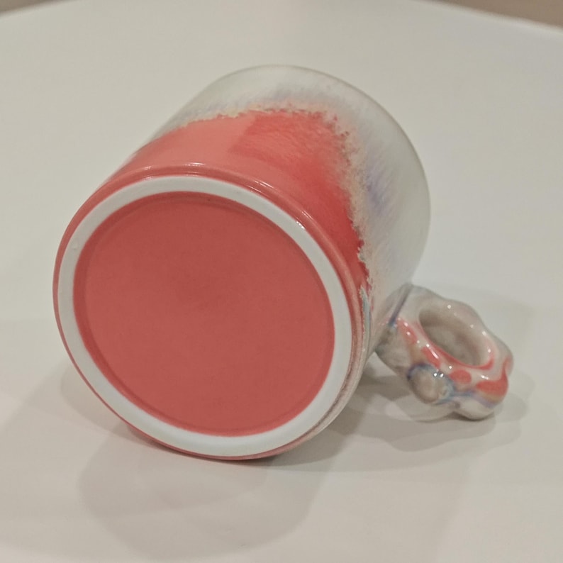 May include: A ceramic mug with a coral-colored base and a white rim. The exterior transitions from coral to white with hints of blue. The handle is a unique, rounded shape. This mug is ideal for coffee, tea, or other beverages.