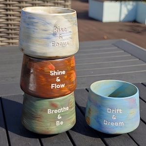 May include: Four stacked ceramic cups with inspirational phrases. The top cup is white and blue with "Rise & Focus". The second cup is brown with "Shine & Flow". The third cup is green with "Breathe & Be". A blue cup with "Drift & Dream" is next to the stack.