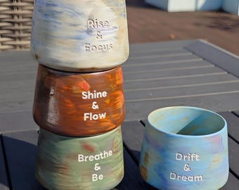 May include: Four ceramic cups with motivational phrases. The top cup is white and blue with "Rise & Focus". The second cup is brown with "Shine & Flow". The third cup is green with "Breathe & Be". A blue cup with "Drift & Dream" is next to the stack.