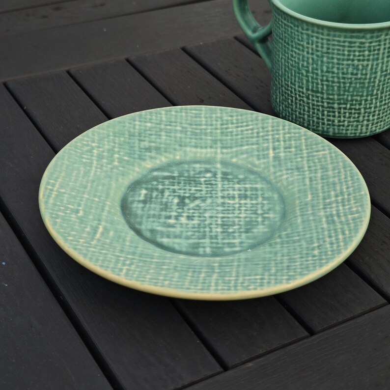 May include: A teal ceramic plate and mug set with a textured woven pattern. The plate is round with a slightly raised center and a cream-colored rim. The mug has a handle and matches the plate's design.