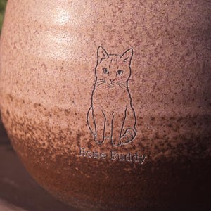 Może przedstawiać: Okrągły ceramiczny kubek z nakrapianym wykończeniem typu gradient, przechodzącym od jasnego r&oacute;żu u g&oacute;ry do ciemnego brązu u podstawy. Powierzchnia zawiera wygrawerowany rysunek siedzącego kota oraz napis Home Buddy wykonany czcionką szeryfową.