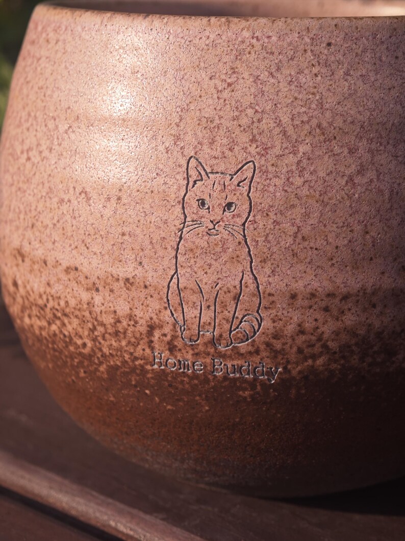 Może przedstawiać: Okrągły ceramiczny kubek z nakrapianym wykończeniem typu gradient, przechodzącym od jasnego r&oacute;żu u g&oacute;ry do ciemnego brązu u podstawy. Powierzchnia zawiera wygrawerowany rysunek siedzącego kota oraz napis Home Buddy wykonany czcionką szeryfową.
