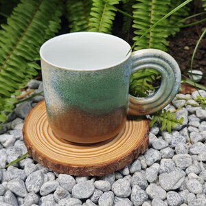 K&ouml;nnte beinhalten: Eine Keramiktasse mit einem einzigartigen Griffdesign, das einen Farbverlauf von Braun &uuml;ber Gr&uuml;n bis Blau aufweist. Die Tasse steht auf einem Holzuntersetzer, umgeben von kleinen Steinen und Gr&uuml;n.