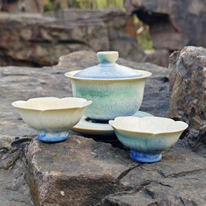 May include: A ceramic tea set with a floral design. The set includes a lidded gaiwan, a saucer, and two small teacups. The gaiwan and teacups have a cream-colored exterior with blue and green accents. The set is displayed on a rock surface.