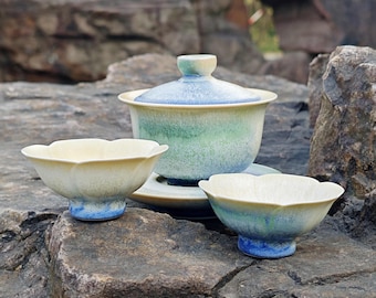 May include: A ceramic tea set with a floral design. The set includes a lidded gaiwan, a saucer, and two small teacups. The gaiwan and teacups have a cream-colored exterior with blue and green accents. The set is displayed on a rock surface.
