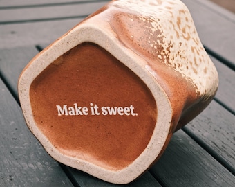 Puede incluir: Una taza de cerámica con una forma única y asimétrica. La taza presenta un exterior moteado de color marrón claro y crema y un interior marrón liso. La frase "Make it sweet" está impresa en blanco en la base de la taza.