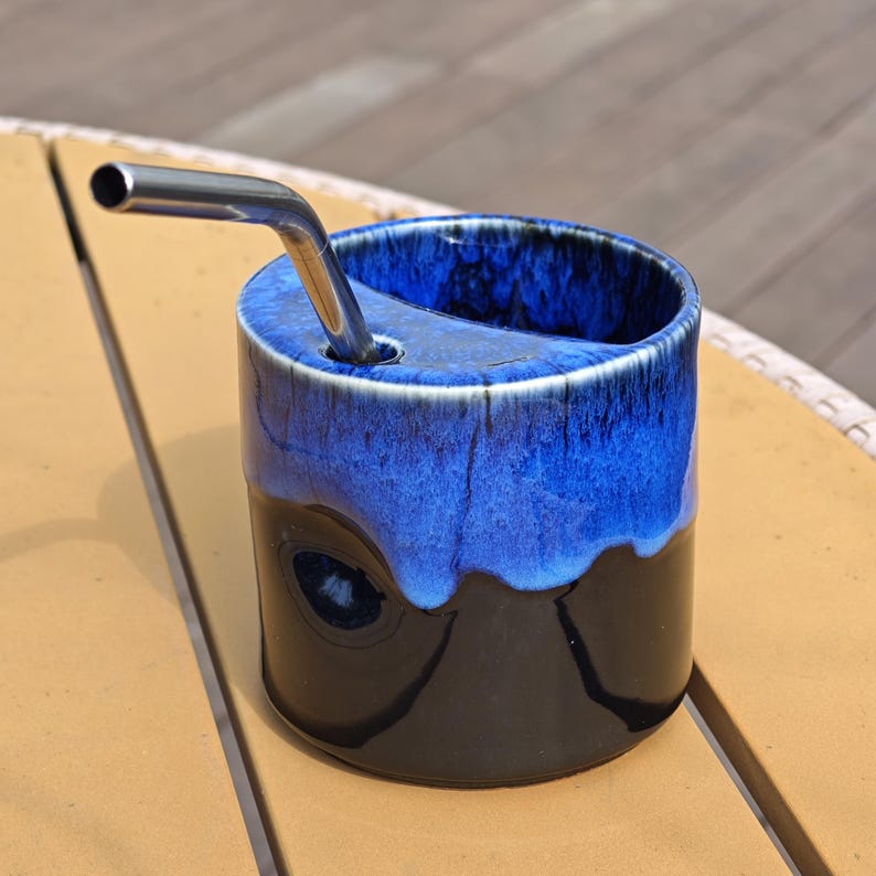 May include: A cylindrical ceramic cup featuring a glossy black base and a vibrant blue drip glaze pattern on the upper portion. The cup includes a built-in circular opening on the top rim, which holds a metallic silver-colored drinking straw.