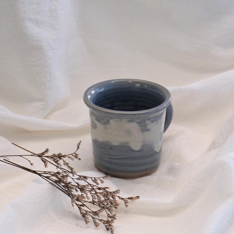 May include: A handcrafted ceramic mug with a blue and white glaze. The mug has a cylindrical form with a small handle and a slightly flared rim. The color scheme is a blend of blues and whites, producing a tranquil, cloudy appearance. Suitable for hot or cold drinks.