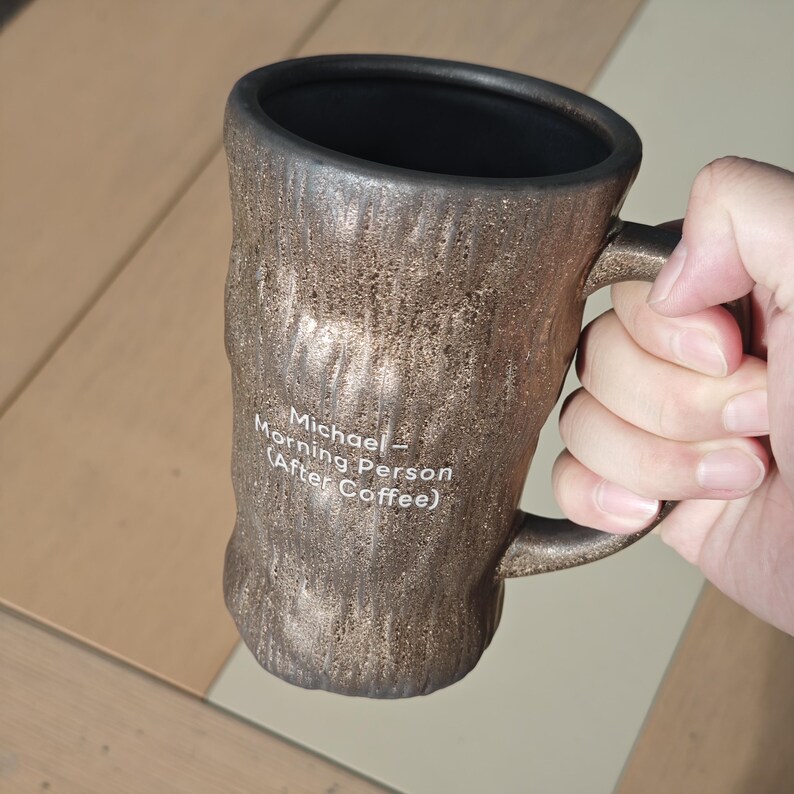 Puede incluir: Una taza cil&iacute;ndrica de cer&aacute;mica con un acabado de bronce met&aacute;lico texturizado que se asemeja a la corteza de un &aacute;rbol. El interior es liso y negro. En el lateral, impreso en texto blanco, se lee: Michael&mdash;Morning Person (After Coffee).