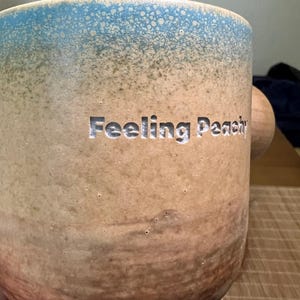 May include: A ceramic mug with a light blue speckled rim and a tan body. The words "Feeling Peachy" are carved into the side. The mug has a wooden handle.