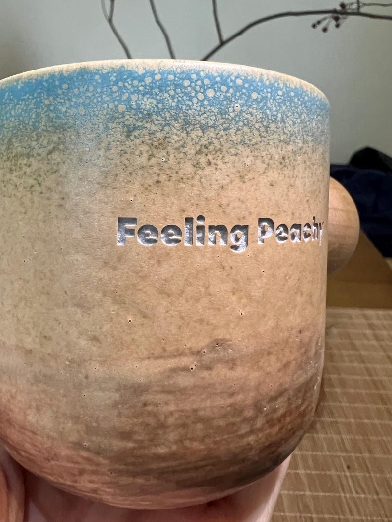 May include: A ceramic mug with a light blue speckled rim and a tan body. The words "Feeling Peachy" are carved into the side. The mug has a wooden handle.