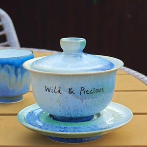 May include: A light blue ceramic tea set with a matching lid and saucer. The tea cup has the words "Wild & Precious" written on the side. The set is on a yellow table.
