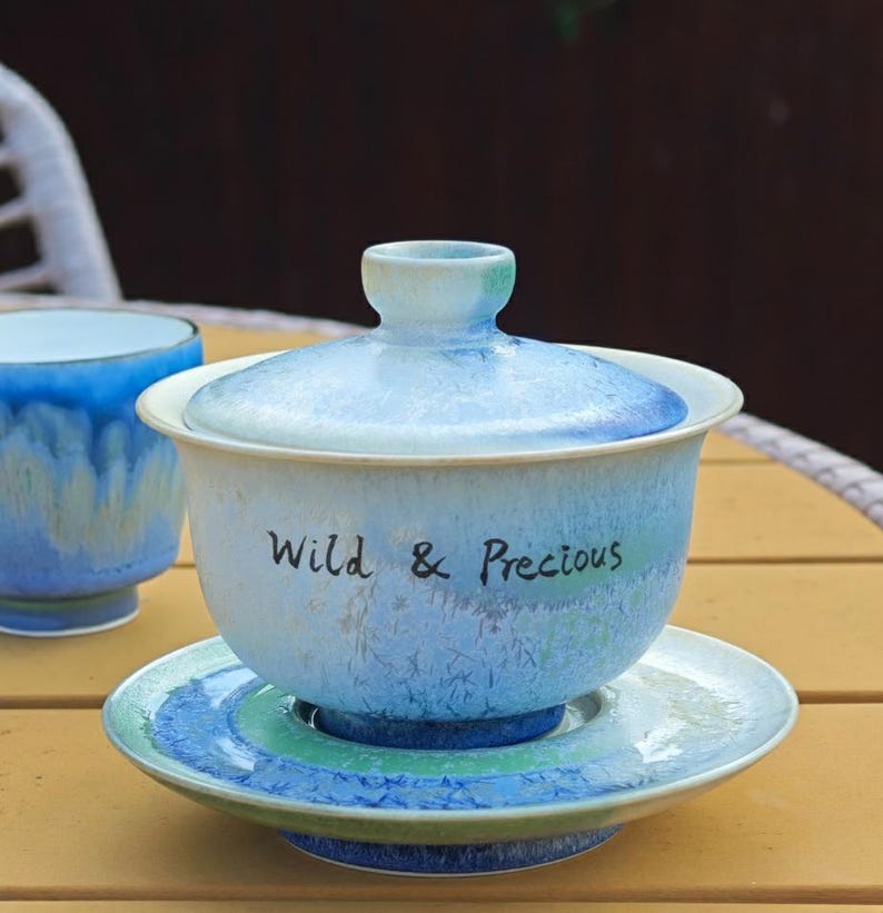 May include: A light blue ceramic tea set with a matching lid and saucer. The tea cup has the words "Wild & Precious" written on the side. The set is on a yellow table.