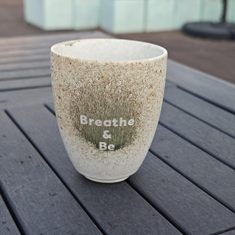 Puede incluir: Un vaso de cer&aacute;mica moteada con las palabras "Breathe & Be" en letras blancas. La taza tiene una base beige claro con un patr&oacute;n moteado y un &aacute;rea verde m&aacute;s oscura detr&aacute;s del texto. El vaso est&aacute; sobre una superficie gris.