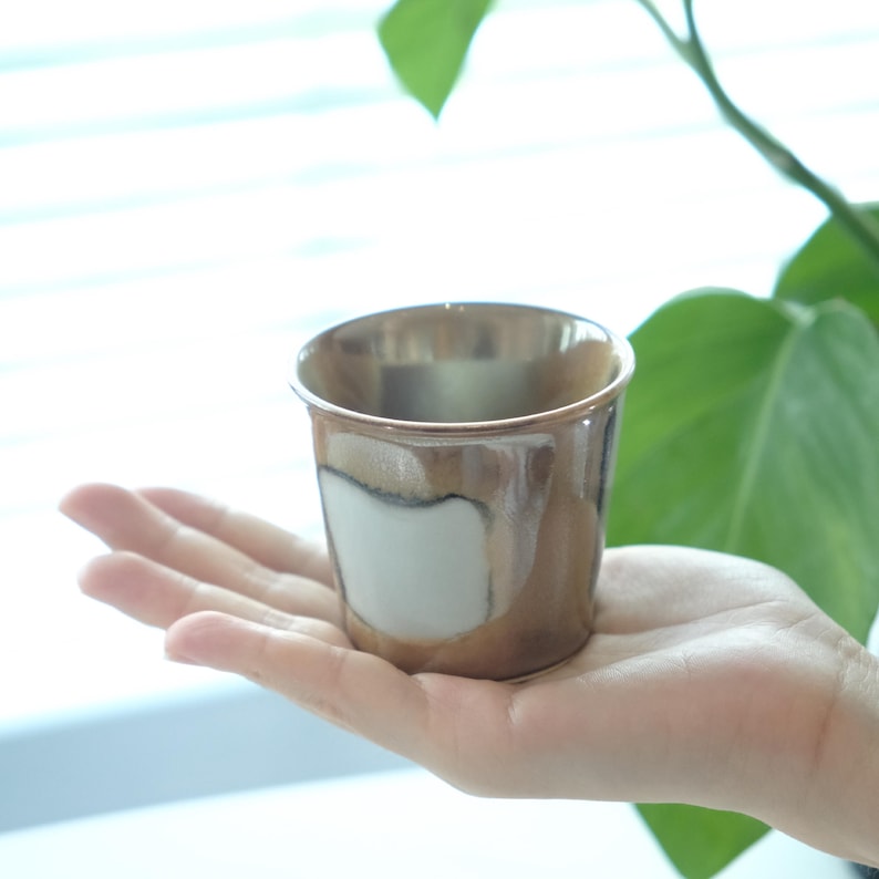 以下が含まれることがあります： メタリックゴールドの縁と茶色と白の釉薬が施された小さな手作りのセラミックカップ。カップは人の手で持たれており、そのユニークなデザインと質感を際立たせています。飲用に使用される可能性が高いです。
