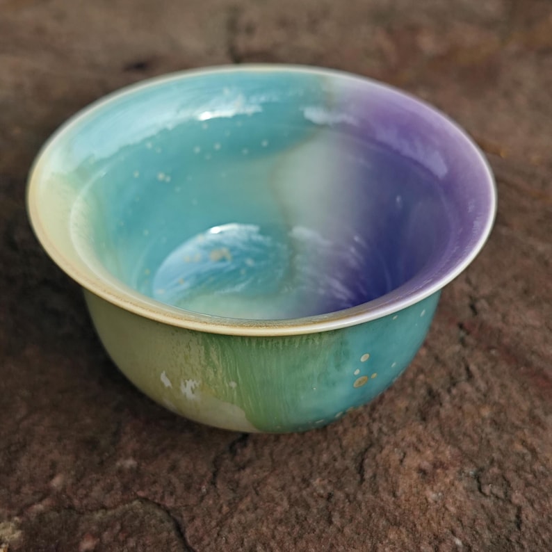 May include: A ceramic bowl with a gradient of colors, including green, blue, and purple. The bowl has a wide rim and a smooth, glossy finish. Small gold-colored speckles are scattered across the surface. The bowl is likely used for serving or display.
