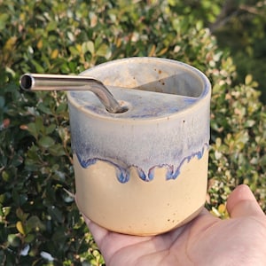 Puede incluir: Un vaso de cerámica hecho a mano con una pajita de acero inoxidable. El vaso tiene una base beige claro con una parte superior esmaltada azul y blanca. La pajita se inserta en un agujero en la parte superior del vaso.
