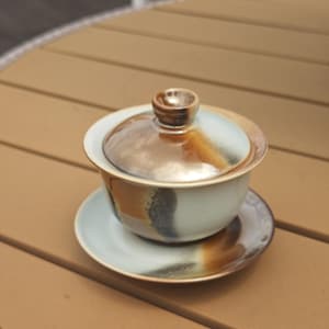 May include: A ceramic gaiwan tea set consisting of a bowl, a lid with a small knob, and a saucer. The set features a light blue base glaze with blended brown and dark grey accents that create a gradient effect across the surface of each piece.