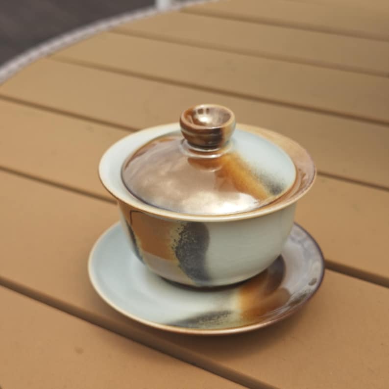 May include: A ceramic gaiwan tea set consisting of a bowl, a lid with a small knob, and a saucer. The set features a light blue base glaze with blended brown and dark grey accents that create a gradient effect across the surface of each piece.