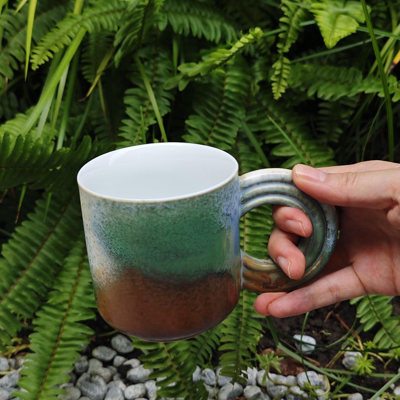 K&ouml;nnte beinhalten: Eine Keramik-Tasse mit Farbverlauf in Gr&uuml;n, Blau und Braun. Die Tasse hat einen wei&szlig;en Innenraum und einen gro&szlig;en, abgerundeten Henkel. Die Tasse wird in einer Hand gehalten, mit gr&uuml;nem Laub im Hintergrund.