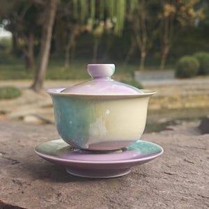 May include: A ceramic tea set with a lid, cup, and saucer. The cup and saucer are a gradient of colors, including light blue, yellow, and purple. The tea set is sitting on a stone surface.