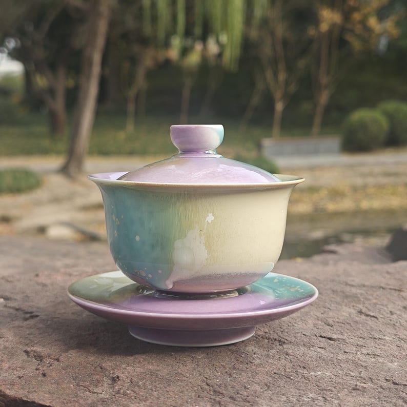 May include: A ceramic tea set with a lid, cup, and saucer. The cup and saucer are a gradient of colors, including light blue, yellow, and purple. The tea set is sitting on a stone surface.