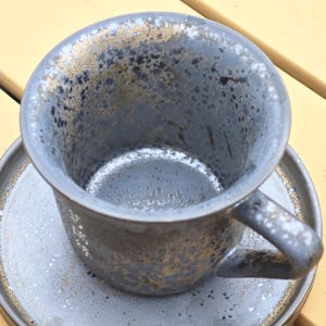 May include: A handmade ceramic coffee cup and saucer set. The cup and saucer are a dark gray color with a speckled texture and gold accents around the rim and edges. The cup has a curved handle.