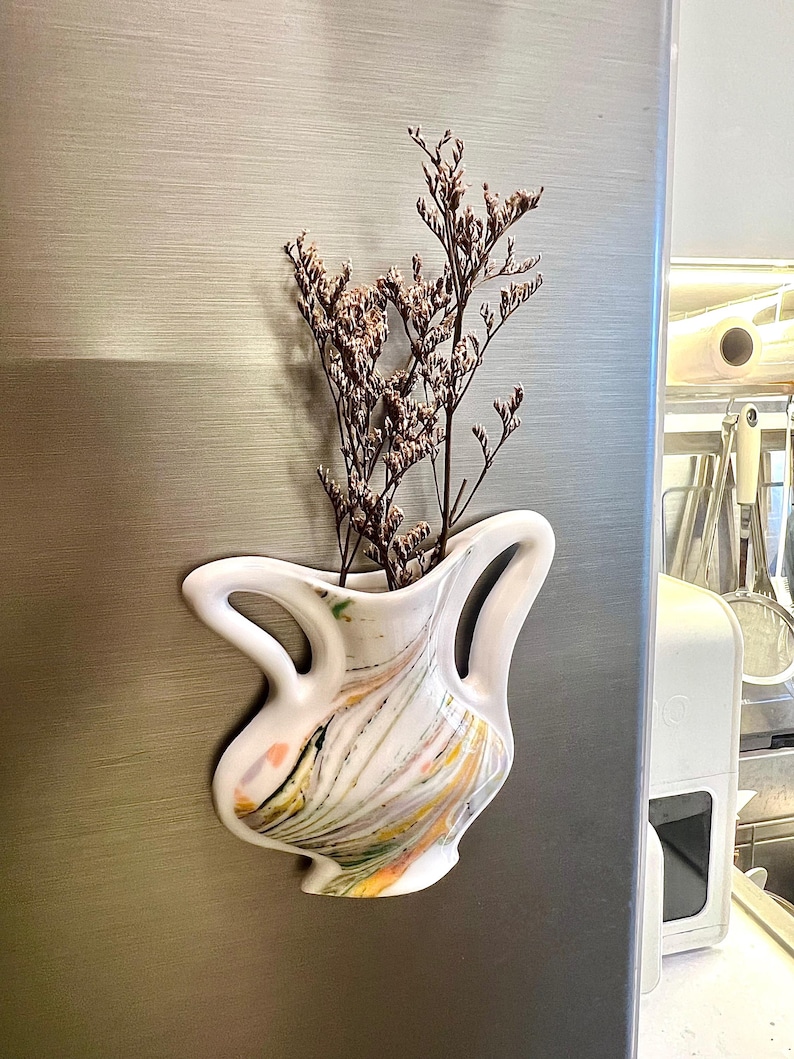 Op de afbeelding: Een witte keramische vaas met een uniek, gebogen handvatontwerp, versierd met kleurrijke abstracte verfstreken. De vaas is gevuld met gedroogde, bruine bloemstelen, wat een decoratieve weergave cre&euml;ert. De vaas is bevestigd aan een metalen oppervlak.