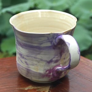 May include: A handmade ceramic mug with a unique marbled design in shades of purple and white. The mug has a rounded body, a sturdy handle, and a slightly irregular rim. The mug is set on a wooden surface.